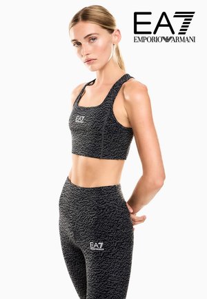 Frau trägt schwarzen, gemusterten Sport-BH und Leggings mit EA7 Emporio Armani-Logos, posiert vor weißem Hintergrund.