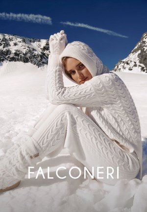 Donna con felpa bianca a trecce con cappuccio, pantaloni abbinati e guanti seduta sulla neve con montagne e cielo azzurro limpido sullo sfondo.