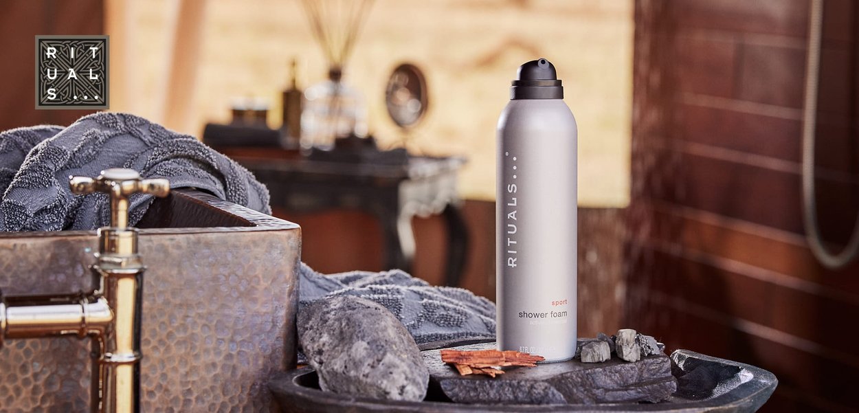 Silver cylindrical spray can with black cap labeled "RITUALS sport shower foam" placed on a dark wooden tray with grey stones and wooden accents.