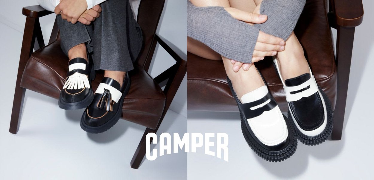 Dos pares de pies con mocasines Camper en blanco y negro, sentados en sillas de cuero marrón, con las piernas cruzadas y las manos apoyadas en las rodillas.