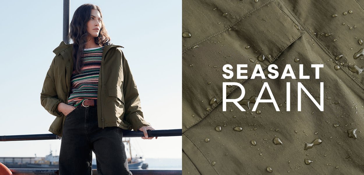 Olive green waterproof jacket featuring a zip, hood, and textured fabric with water droplets. Styled with a striped top and black jeans.
