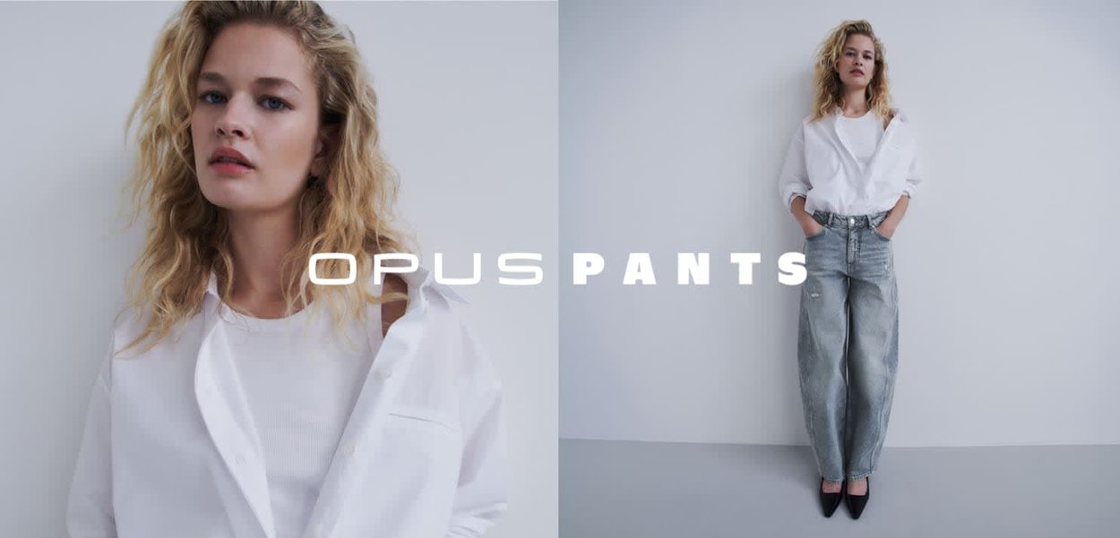 Blonde Frau mit welligem Haar, die ein weißes Hemd und lockere graue Jeans trägt, steht vor einem schlichten hellgrauen Hintergrund.