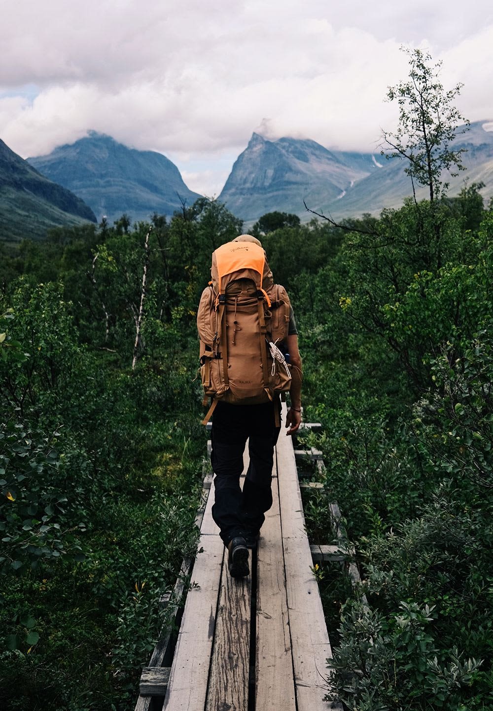 Brun, texturerad ryggsäck med orange topp, buren av en person på en träspång. Lush grön växtlighet och berg i bakgrunden.