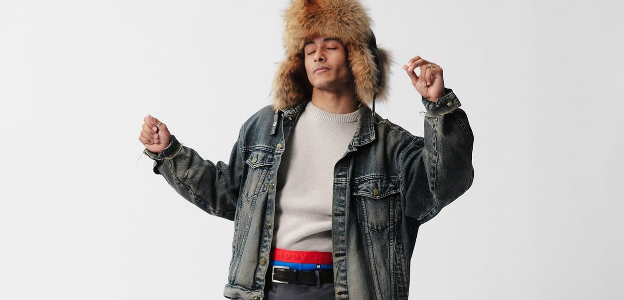 Jeansjacke mit einer Kapuze, die mit Kunstpelz gefüttert ist, hellgrauer Pullover darunter, marineblauer und roter Gürtel sowie graue Hose. Neutraler Hintergrund.