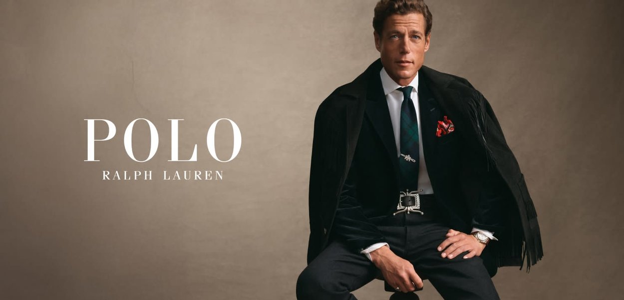 Blazer en velours noir avec des franges, chemise blanche, cravate à carreaux, boucle de ceinture en argent. Une poche carré rouge ajoute du contraste. Fond neutre.