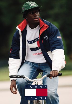Jeune homme portant une casquette verte, une veste Tommy Jeans bleu marine et blanche, un sweat-shirt blanc avec logo, et un jean, assis sur un vélo en plein air.