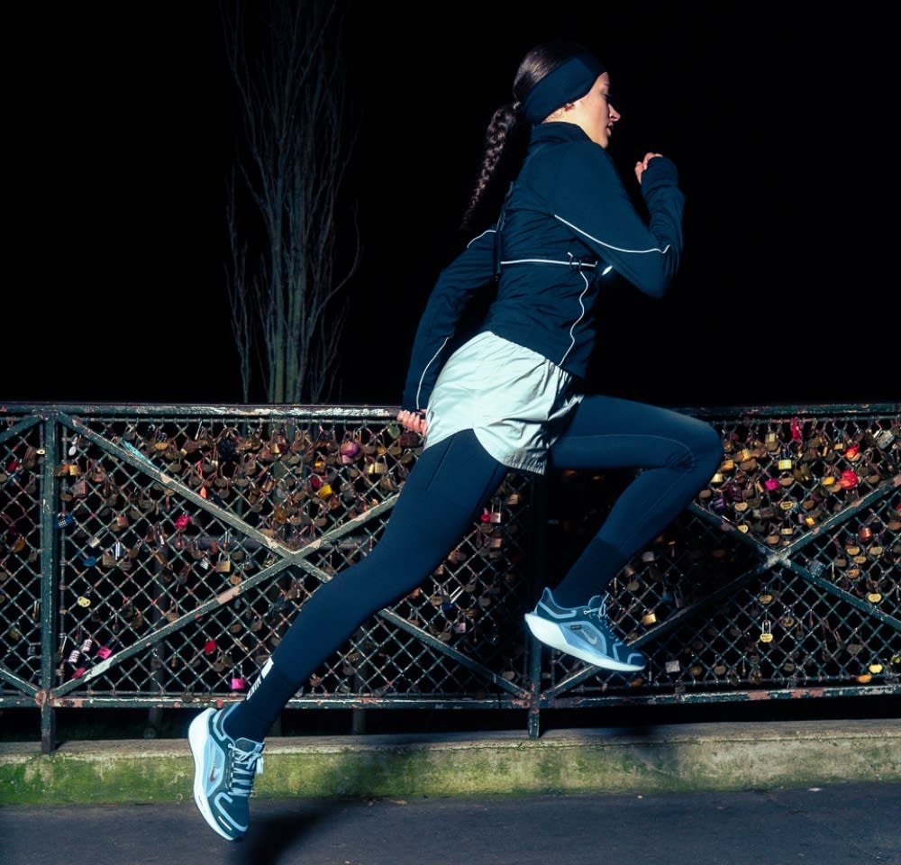 Athletic footwear and attire: black long sleeve top, light gray shorts, black leggings, and gray sneakers, in a dynamic running pose against a dark background.
