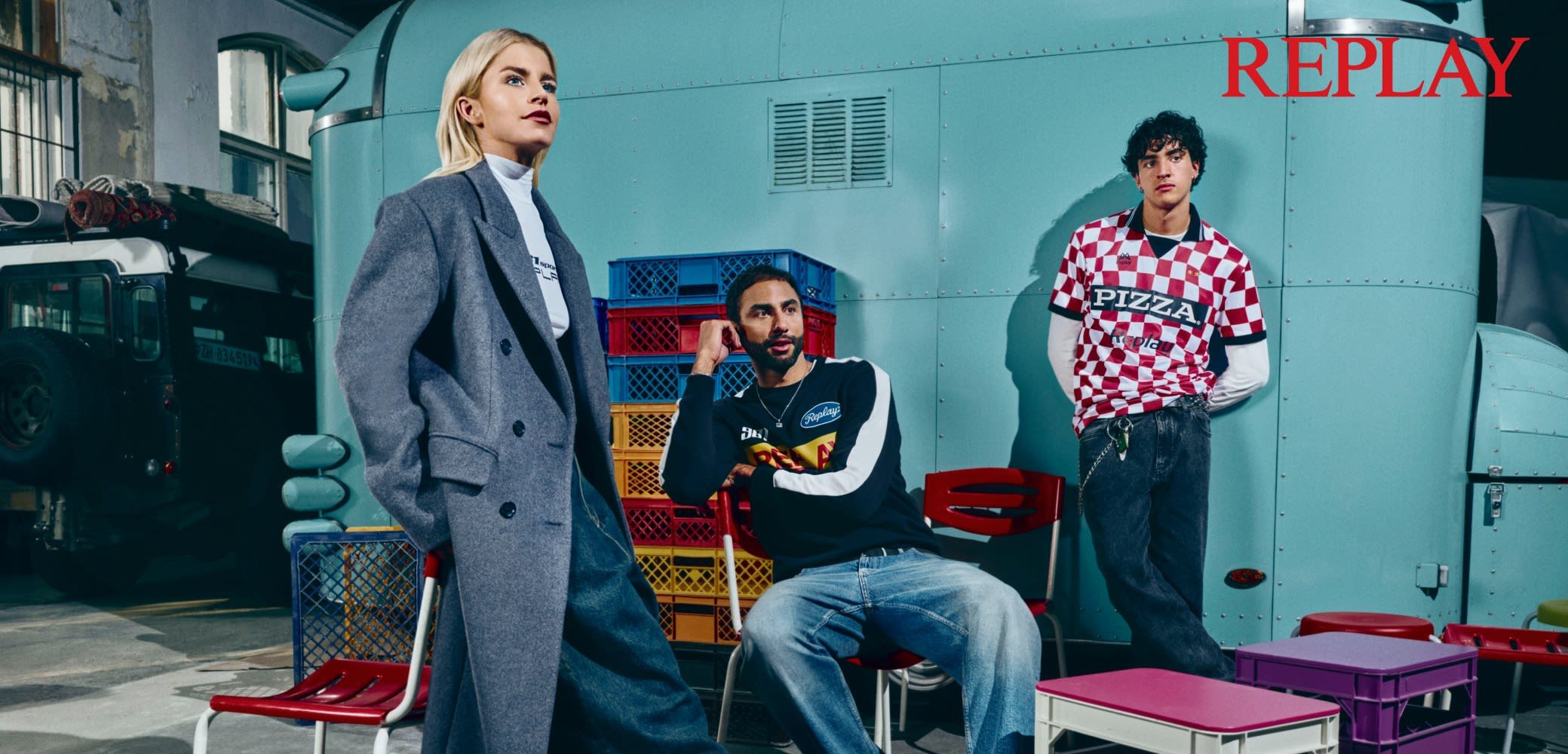 Tre persone posano in un ambiente colorato. Indossano un cappotto grigio, una felpa nera con un logo e una camicia a quadri rossa con la scritta "PIZZA". Crate e sedie colorate arricchiscono lo sfondo.