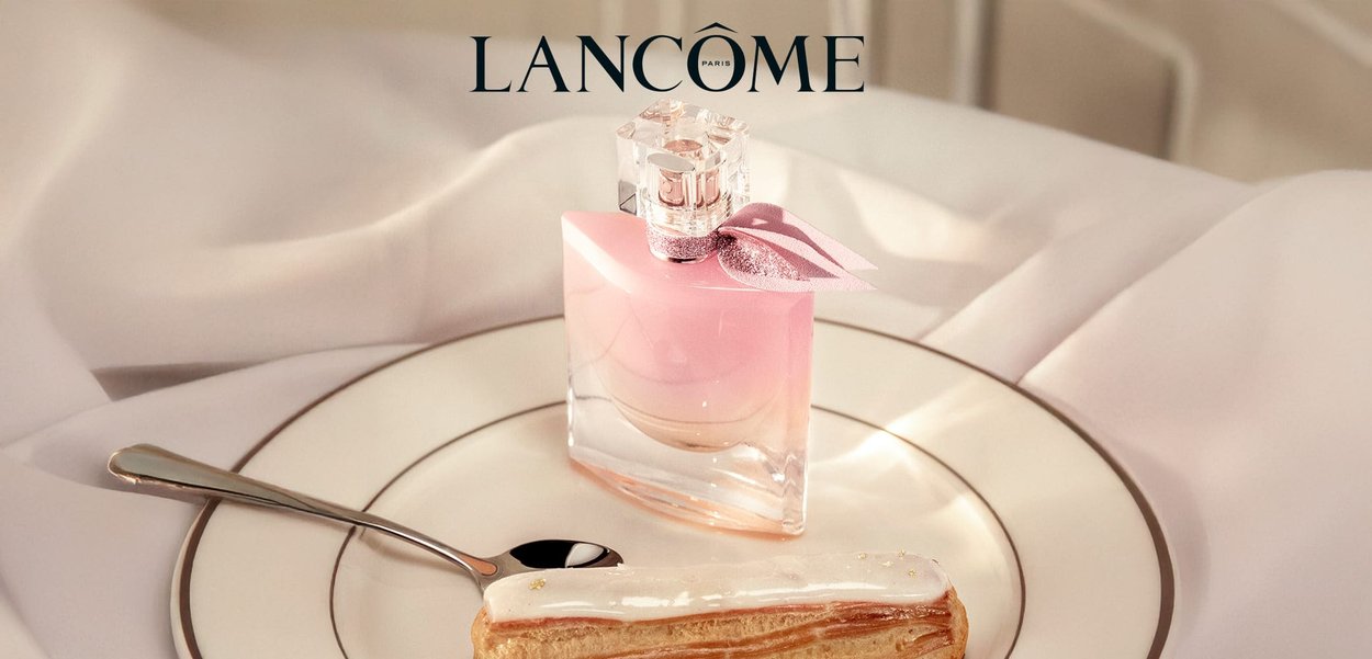 Pink glass perfume bottle with a clear square cap and a pink ribbon, sitting on a plate with a cream-filled eclair and silver spoon.