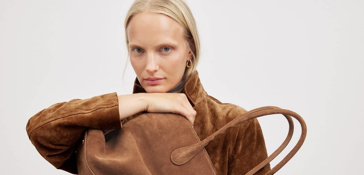 Brązowa zamszowa torba typu tote o miękkiej fakturze i zaokrąglonych uchwytach, trzymana przez osobę ubraną w brązową zamszową kurtkę. Neutralne tło.