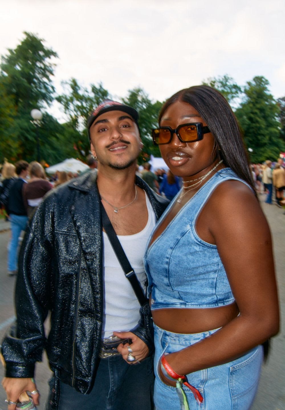 Två personer poserar tillsammans. Den ena har en glänsande svart jacka över en vit skjorta, den andra har en denimcroppad topp och matchande shorts.