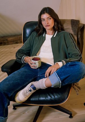 Veste bomber verte avec accents blancs, haut crème, jean en denim et baskets. Assis sur une chaise noire, tenant une tasse en céramique.