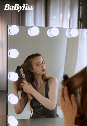 En kvinne bruker en rund hårbørste med metallisk sylinder foran et opplyst sminkespeil, iført en grå topp og sølvkjede.