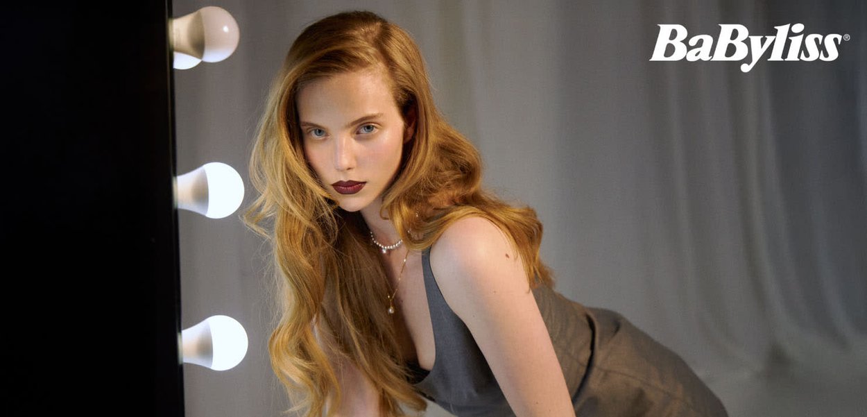 Grå antrekk, bølgete blondt hår, minimal sminke med mørke lepper, innrammet av lyse pærer og mykt, bølgende stoffbakgrunn. BaByliss-logo til stede.