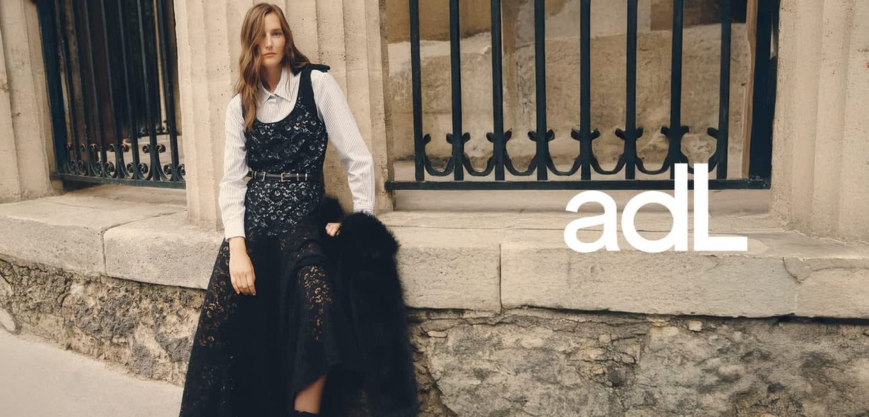 Zwarte kanten jurk over een wit gestreept shirt, ceintuur in de taille. Zwarte bontstola over de schouder gedrapeerd. Achtergrond met stenen muur en ijzeren tralies.