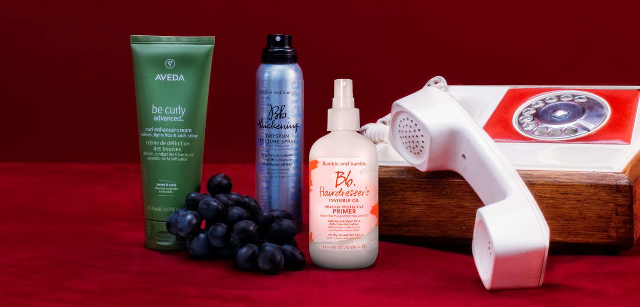 Grüne Aveda Lockenverstärker-Creme, blauer Bumble and Bumble Spray, weiße und orange Haarprimerflasche und ein vintage rotes Wählscheibentelefon auf einer roten Fläche.