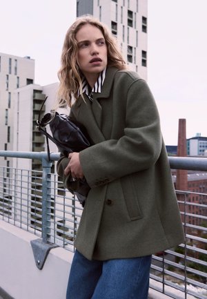 Olive green wool overcoat with a double-breasted design, worn over a black and white striped shirt, paired with blue denim and a black bag.