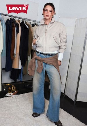 Junge Frau steht in einem Raum mit Kleidergestell, trägt eine beige Bomberjacke, Jeans mit weitem Bein und ein Sweatshirt, das um die Taille gebunden ist, darüber das Levi’s-Logo.