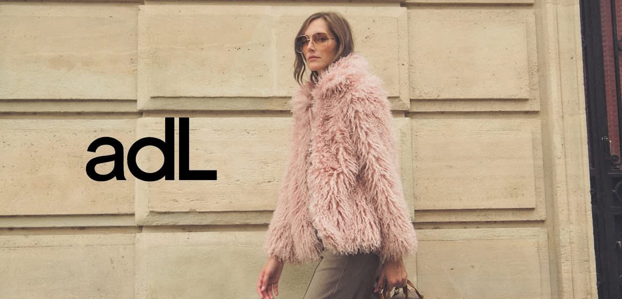 Roze pluizige bontjas met een hoge kraag, gedragen over een grijze outfit, gecombineerd met ronde zonnebril en een handtas, tegen een stenen muur.