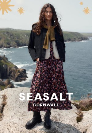 Schwarze Jeansjacke über einem grauen Pullover, Maxi-Kleid mit floralem Muster in dunklen Farben und schwarze Stiefeletten, vor einer Küstenlandschaft.