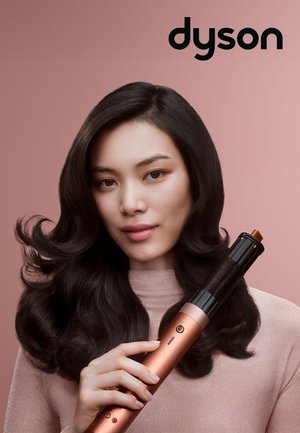 Frau mit langen, dunklen, welligen Haaren hält einen roségoldenen Lockenstab vor einem rosa Hintergrund, darüber das Markenlogo „dyson“.