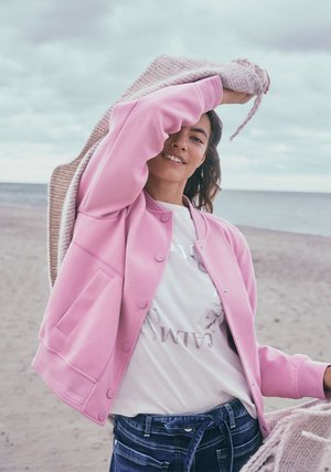 Sweatshirt rose avec des boutons, col rond, associé à un t-shirt graphique blanc et un jean foncé. Le modèle tient une couverture légère sur une plage.