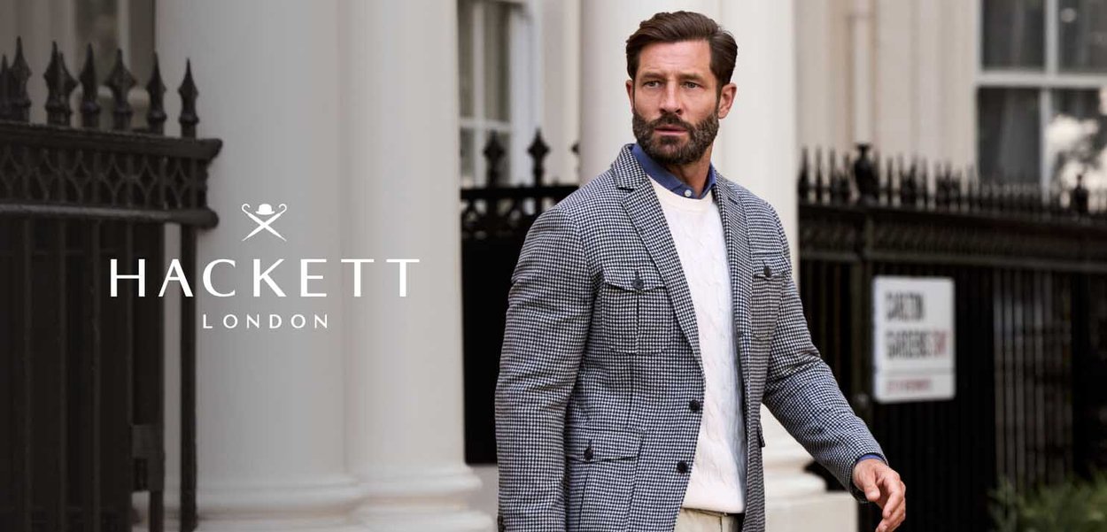 Blazer gris de pata de gallo sobre un suéter blanco de punto trenzado, combinado con pantalones beige; con un fondo urbano clásico.