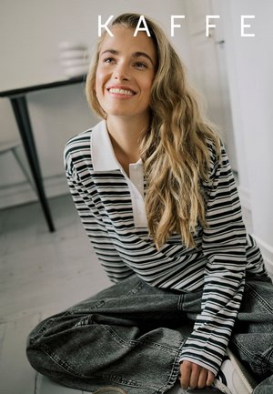 Junge Frau mit langen, welligen Haaren, die ein gestreiftes Shirt und Jeans trägt, sitzt auf dem Boden und lächelt in einem minimalistischen Raum.