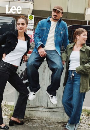 Trois personnes posent à l'extérieur. L'une porte une veste en denim noire et un pantalon noir, une autre une veste en denim bleue, et la dernière une veste olive.