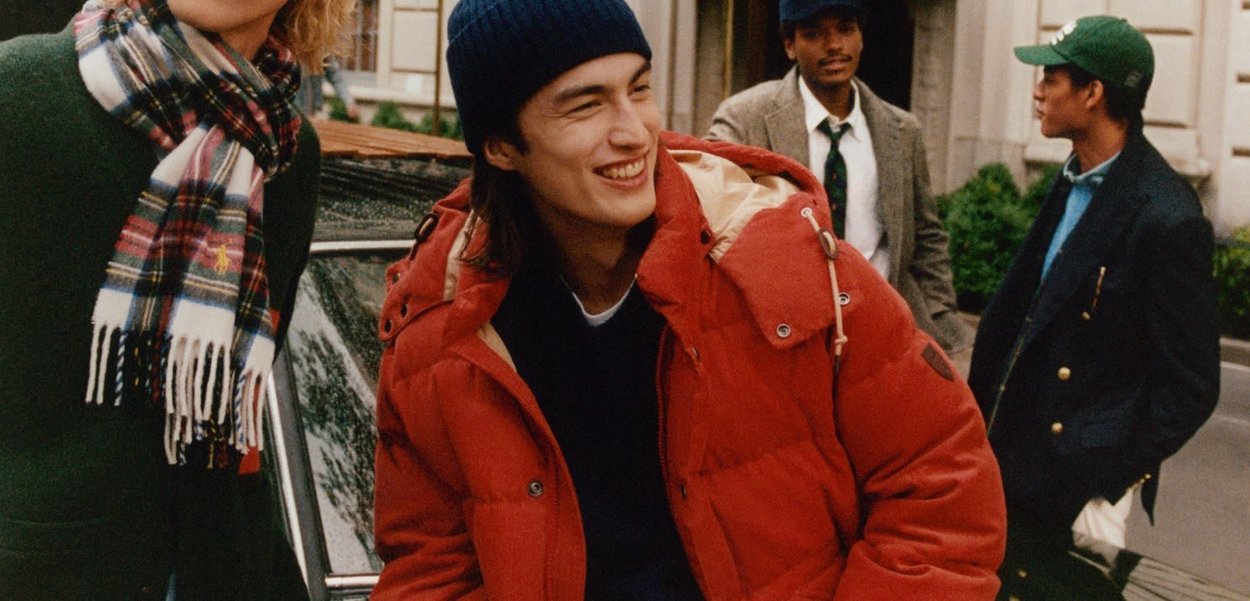 Red puffer jacket with a hood, worn over a black sweater. The scarf features a plaid pattern. Background includes a car and three individuals.