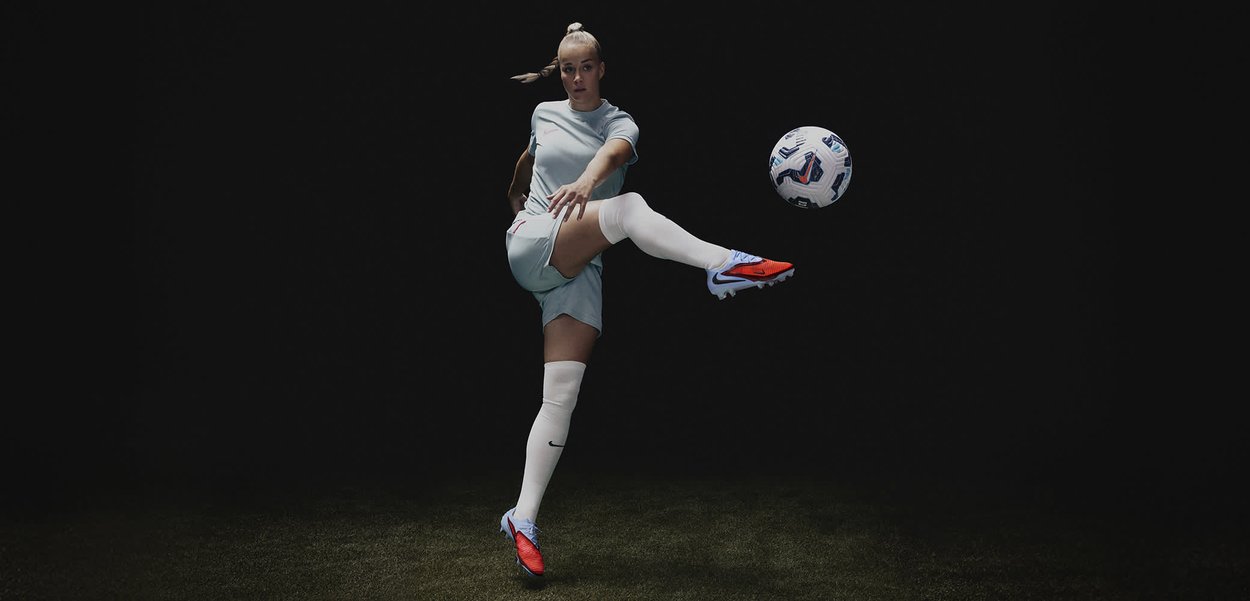Giocatore di calcio in uniforme azzurro chiaro con scarpe da calcio rosso e blu, che calcia un pallone bianco con motivo blu, su uno sfondo nero.
