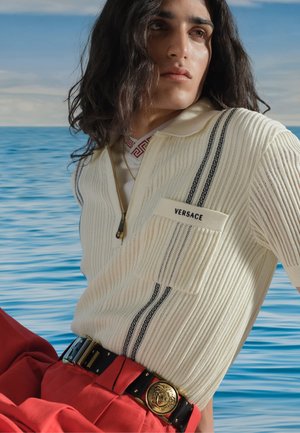 Giovane con capelli lunghi e scuri indossa un top in maglia crema Versace e pantaloni rossi con cintura nera, posa vicino al mare blu calmo sotto un cielo nuvoloso.