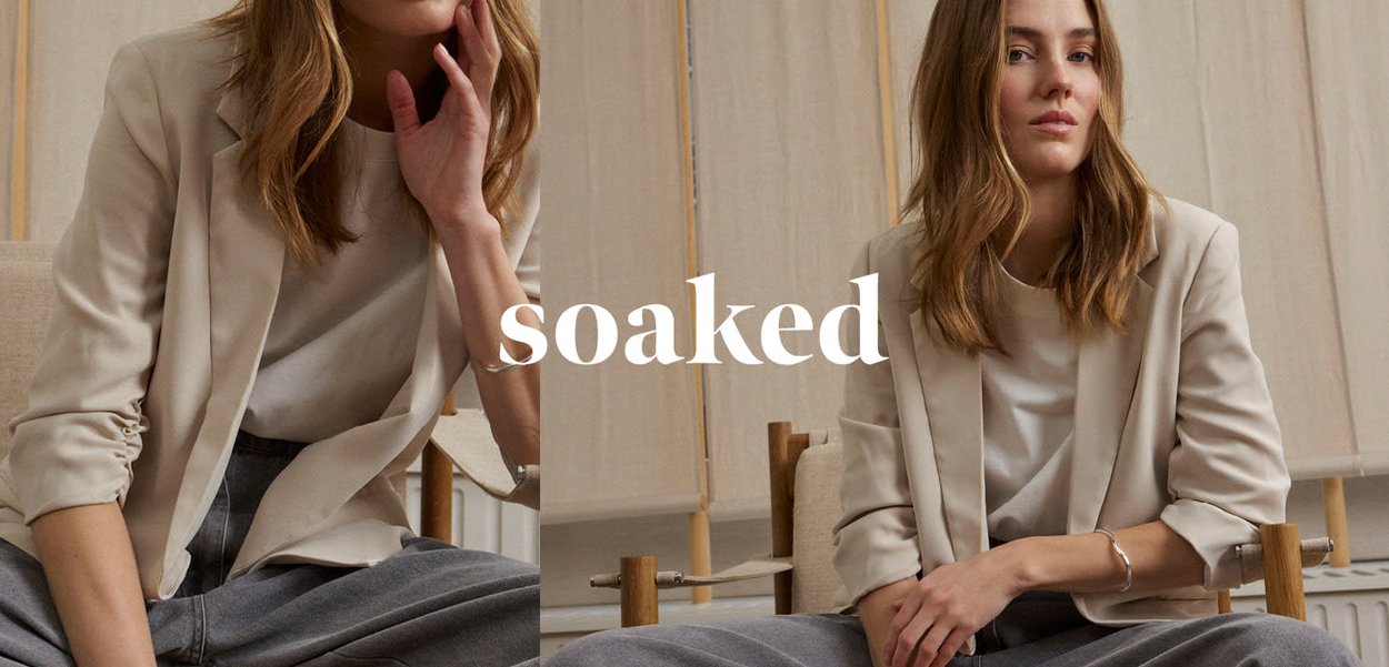Junge Frau in lässigem beigem Blazer und grauer Hose sitzt selbstbewusst in einem Raum mit neutralen Farbtönen, darüber der Schriftzug "soaked".