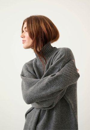 Pull en tricot gris avec un col côtelé haut, des épaules tombantes et des manches longues. Texture douce et coupe décontractée. Modèle de profil.