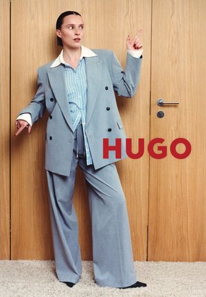 Donna con abito azzurro chiaro e camicia a righe in piedi davanti a porte di legno, indica verso l'alto con entrambe le mani, testo rosso "HUGO" vicino.