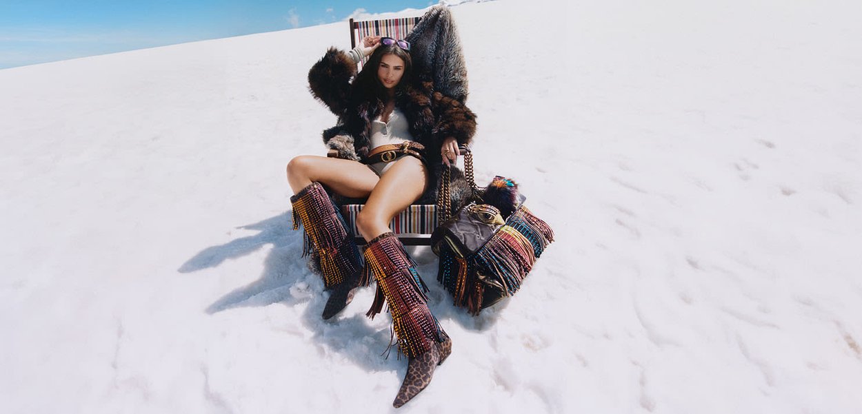 Femme portant un manteau en fourrure, des bottes à franges et des lunettes de soleil, assise sur une chaise dans un paysage enneigé avec un sac à franges à côté d'elle.