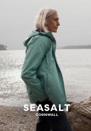 Kvinde med langt blondt hår iført en teal hættetrøje står ved en rolig, overskyet kyst med stenet strand og fjerntliggende land.