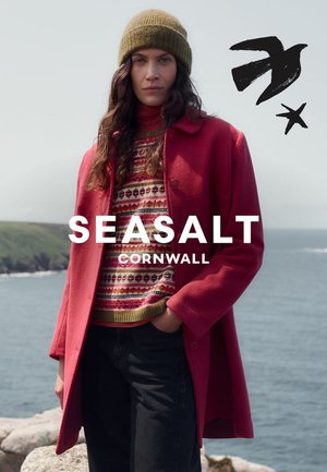 Roter, strukturierter Mantel über einem mehrfarbig gemusterten Pullover, kombiniert mit schwarzen Jeans. Das Model trägt eine olivgrüne, gestrickte Mütze als Accessoire.