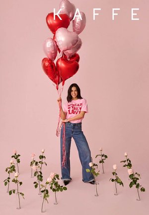 Junge Frau hält rote und pinkfarbene herzförmige Luftballons und steht zwischen pinkfarbenen Rosen in Glassvasen vor einem pinken Hintergrund mit dem Schriftzug "KAFFE".