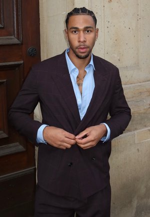 Un costume croisé de couleur bordeaux foncé avec une chemise habillée bleue en dessous, présentant un motif à carreaux et une texture lisse. Des mains ajustent la veste.