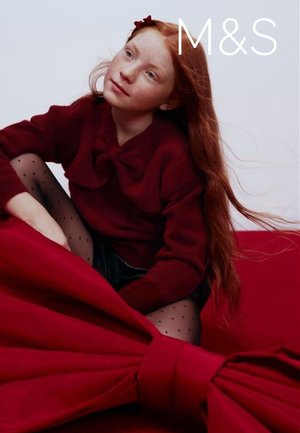 Rode gebreide trui met strikdetail, zwarte korte broek en panty's met polkadots. Het model zit op een rode stoffen achtergrond. Lang rood haar.