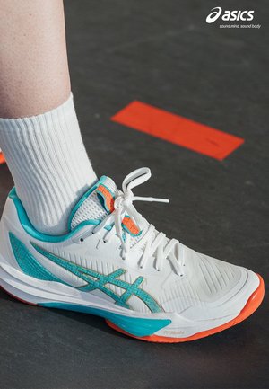 Hvid og turkis ASICS løbesko med orange sål og detaljer, båret med hvide sokker på en mørk overflade med røde markeringer.