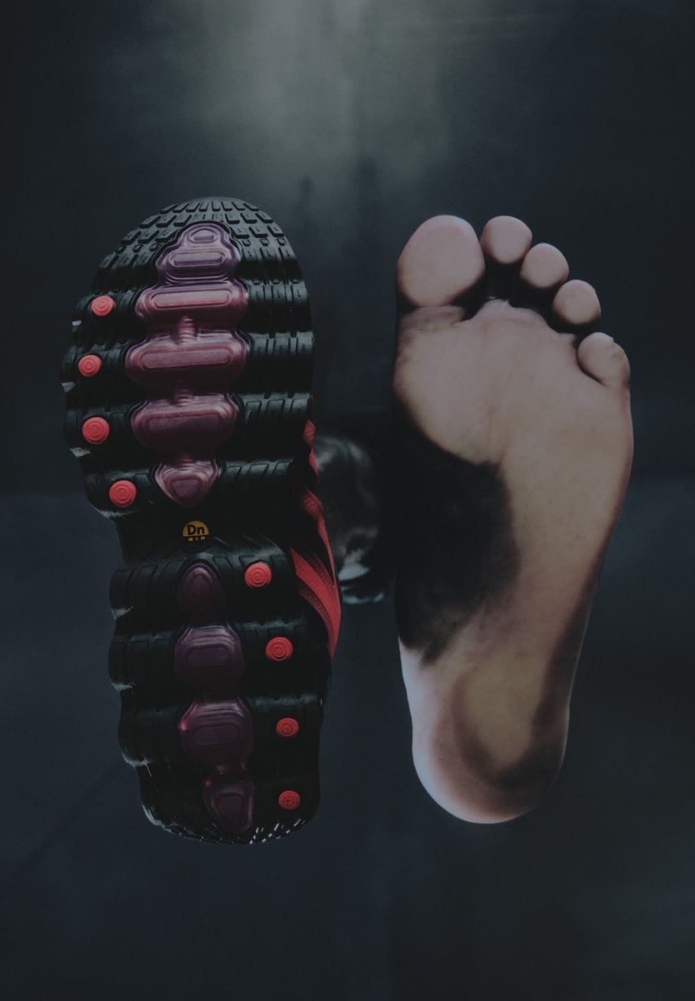 Zwarte sneaker met paarse en rode accenten, met een getextureerde zool, naast een blote voet geplaatst, waardoor de vorm en details worden benadrukt.