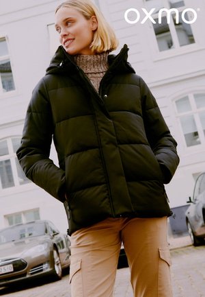 Sort polstret jakke med hætte, med markante vandrette syninger, en frontlynlås og sidelommer, båret over en brun sweater.