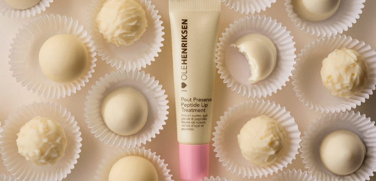 Traitement aux peptides pour les lèvres Pout Preserve dans un tube crème avec des accents roses, entouré de confiseries blanches dans des coupelles en papier à côtes.