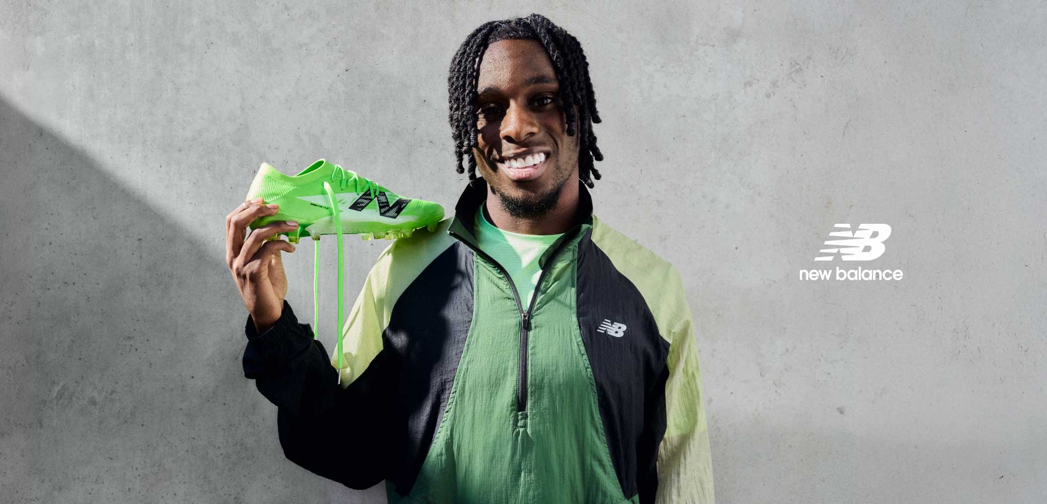 Botte de football vert vif avec un dessus texturé, des accents noirs et des crampons saillants. Le modèle porte une veste multicolore verte, souriant.