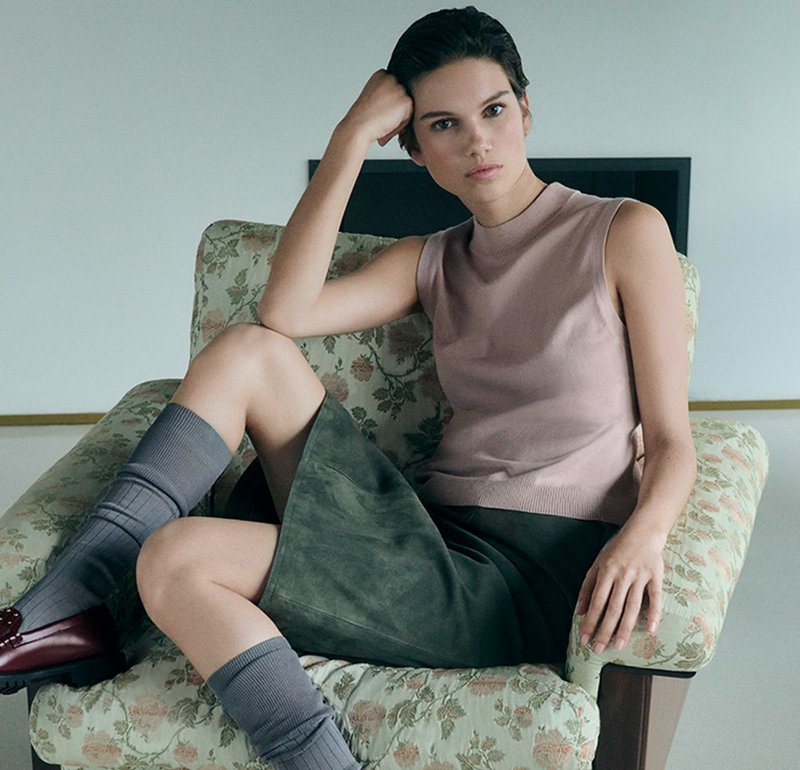 Light pink sleeveless sweater, dark green suede skirt, gray knee-high socks, seated on a floral-patterned armchair with a plain backdrop.