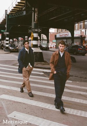 Due modelli maschili camminano su un attraversamento pedonale. Uno indossa un cappotto scuro e un maglione blu navy; l'altro sfoggia un cappotto marrone sopra un cardigan blu navy.