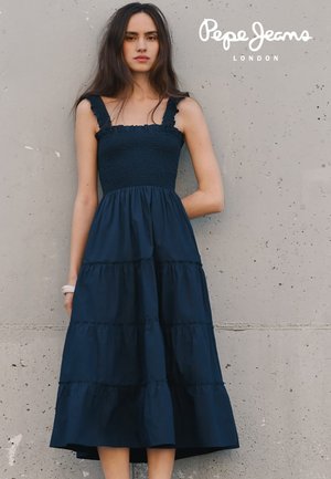 Mujer con un vestido midi sin mangas azul marino de capas con tirantes con volantes, de pie frente a una pared de hormigón texturizada.