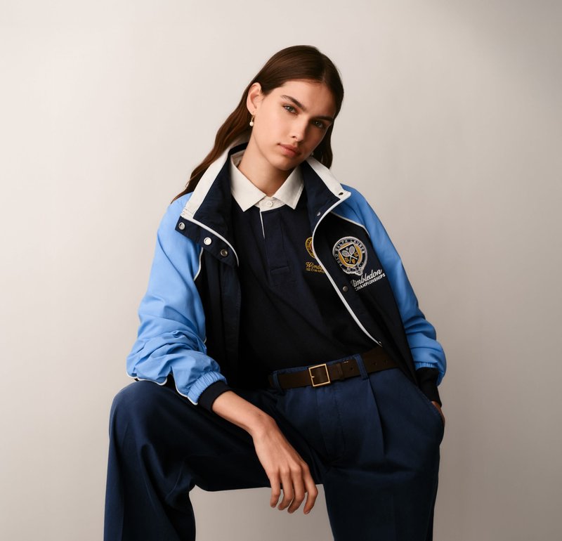 Joven mujer que lleva una chaqueta Wimbledon azul y blanca sobre una camisa polo azul marino y pantalones azules, posando contra una pared clara y lisa.